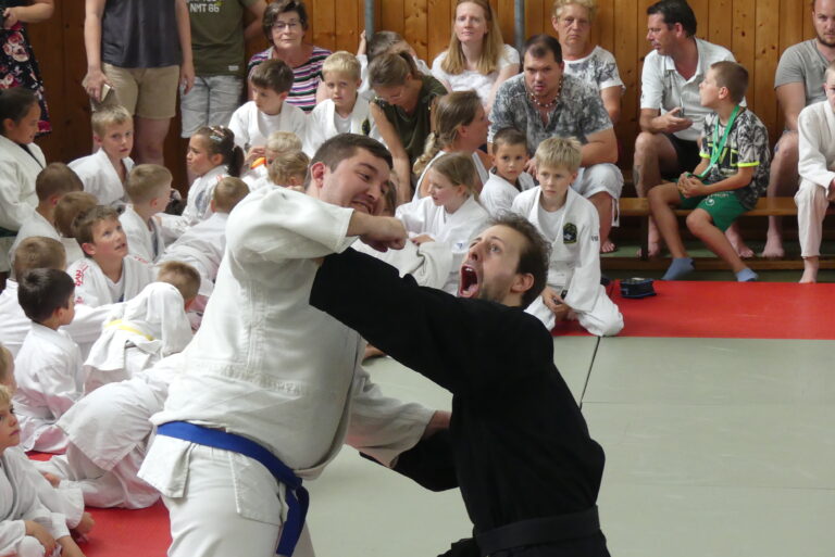 Mehrere Eltern und Kinder auf Bänken sitzend. im Vordergrund kämpfen Moritz Vuckovic und Markus Gstöttner. Moritz trägt einen weißen Gi mit blauem Obi. Seine rechte Hand ist abgewinkelt und zu einer Faust geballt. Markus trägt einen schwarzen Gi. Mit seiner linken Arm wehrt er in ausgestreckter Haltung die Faust ab und schreit mit weit geöffneten Mund.