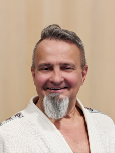 Ein Porträtfoto von Harald Winter, einem Sportler bei JJGE. Er trägt einen weißen Gi und man kann teilweise seine Brust sehen. Er hat seitlich kurzes braun/weißes Haar, am Oberkopf hat er eine leichte Welle. Er trägt einen Goatee-Bart, er ist buschig weiß und in der Mitte ist ein schmaler dunkler Streifen zu sehen. Der Rest des Gesichts ist glatt rasiert. Seine Gesichtsform ist rund und man sieht weiß/schwarze Brusthaare. Er schaut direkt in die Kamera und lächelt und zeigt etwas seine Zähne. Der Hintergrund ist hellbraun