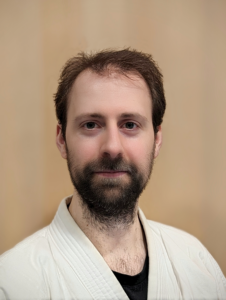 Markus Gstöttner, ein Sportler bei JJGE, ist auf dem Porträtfoto zu sehen. Er trägt einen weißen Gi und darunter ein schwarzes T-Shirt. Sein kurzes dunkelbraunes Haar ist gepflegt und er hat einen dunklen, kurz gestutzten Vollbart. Seine Augen sind blaugrau und er hat einen hohen Stirnansatz. Er schaut direkt in die Kamera und lächelt leicht. Seine Gesichtsform ist länglich und er hat leichte Brusthaare, die oberhalb des T-Shirts leicht zu erkennen sind. Der Hintergrund des Bildes ist hellbraun.