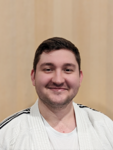 Ein Porträtfoto von Moritz Vuckovic, einem Sportler bei JJGE. Er trägt einen weißen Gi mit schwarzen Streifen auf den Schultern und darunter ein weißes T-Shirt. Er hat kurzes braunes Haar, das leicht nach oben gestylt ist. Er trägt einen leichten Oberlippenbart und einen gleichmäßigen Bartansatz an den Wangen und am Kinn. Er schaut direkt in die Kamera und strahlt Freude aus. Seine Gesichtsform ist rund und seine Augen sind braun. Der Hintergrund ist in Hellbraun gehalten.