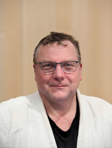 Ein Porträtfoto von Rainer Fritz, dem Obmann Stellvertreter des JJGE. Er trägt eine schlanke schwarze Brille und einen weißen Gi mit einem schwarzen T-Shirt darunter. Ein paar Brusthaare ober halb des T-Shirts sind zu erkennen. Sein kurzes braunes Haar hat leichte graue Strähnen an den Schläfen und er trägt einen leichten Bartansatz an der Oberlippe, den Wangen und dem Kinn. Sein hoher Stirnansatz wird in der Mitte von einigen kurzen Locken verdeckt. Er schaut direkt in die Kamera, lächelt dabei mit geschlossenem Mund und hat eine ovale Gesichtsform mit blaugrauen Augen. Der Hintergrund des Bildes ist hellbraun.