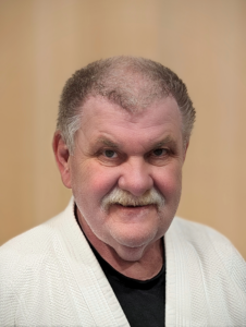 Ein Porträtfoto des Vereinsobmanns des JJGE, Rudolf Zimmerl. Er trägt einen weißen Gi mit einem schwarzen T-Shirt darunter. Sein kurzes Haar ist braun mit einigen grauen Strähnen an den Schläfen. Er hat einen hellbraunen Oberlippenbart in der Mitte seines Gesichts. Sein Stirnansatz ist hoch und hat spitz zulaufende Seiten, während die Mitte seines Gesichts von Haaren bedeckt ist. Er schaut direkt in die Kamera und lächelt. Seine Gesichtsform ist oval und seine Augen sind blaugrau. Der Hintergrund des Bildes ist hellbraun.