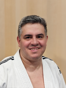 Ein Porträtfoto von Ulrich Inhofner, einem Sportler bei JJGE. Er trägt einen weißen Gi mit blau weißen Streifen auf den Schultern, seine unbehaarte Brust ist leicht zu sehen. Er hat kurz frisiertes braun/weißes Haar, man kann seinen Bartansatz vom Rasieren an der Oberlippe und den Wangen sehen. Er schaut direkt in die Kamera und lächelt und man kann seine Zähne sehen. Seine Gesichtsform ist oval und seine Augen sind braun. Der Hintergrund ist hellbraun.